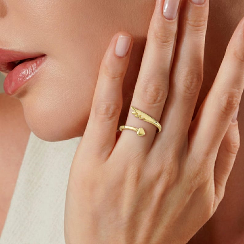 May include: A gold-colored ring with an open design. One end features a wing shape, while the other has a heart-shaped point. The ring is worn on a finger, showcasing its delicate details and elegant style.
