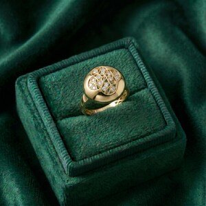 K&ouml;nnte beinhalten: Ein goldener Ring mit einem runden Gesicht, besetzt mit kleinen Diamanten. Der Ring wird in einer gr&uuml;nen Samt-Ringbox pr&auml;sentiert. Das Band des Rings hat ein strukturiertes Design. Das Gesicht des Rings hat ein geometrisches Diamantenmuster.