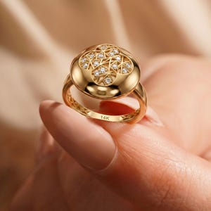 K&ouml;nnte beinhalten: Ein goldener Ring mit gew&ouml;lbtem Oberteil, der ein geometrisches Design und kleine, funkelnde Steine aufweist. Der Ring wird von einer Hand gehalten, die seine komplizierten Details und den 14K-Stempel zeigt.
