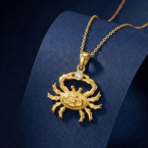 Op de afbeelding: Goudkleurige krabhangernetwerk met een kleine, heldere edelsteen. De krabhanger is gedetailleerd met klauwen en een getextureerd lichaam. De ketting is ook goudkleurig, tegen een donkerblauwe achtergrond.