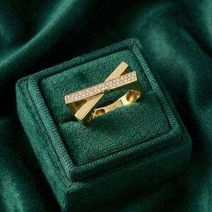 May include: A gold ring with a unique, geometric design. The ring features a band of small, sparkling diamonds set across the top. The ring is displayed in a green velvet ring box, creating a luxurious presentation. The ring is marked "14K".