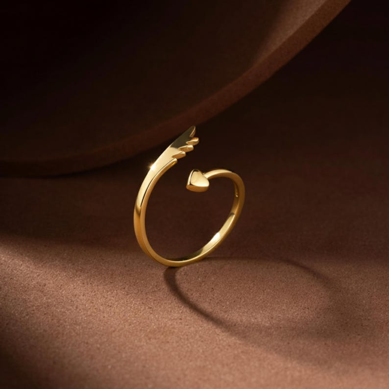 May include: A gold-colored ring with a wing design and a heart-shaped accent. The ring has a delicate, open design, with the wing extending from one side and the heart on the other. The ring is set against a brown background.