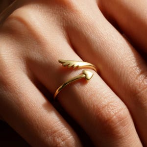 May include: A gold-colored ring with an open design. One end features a wing shape, while the other has a heart. The ring is wrapped around a finger, showcasing its delicate details and shiny finish. The ring is a piece of jewelry.