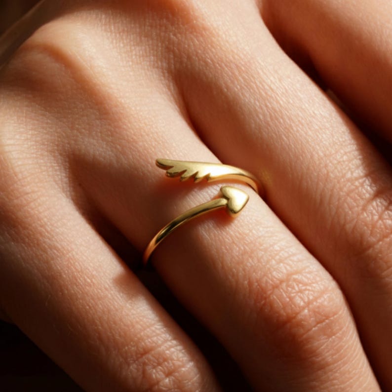 May include: A gold-colored ring with an open design. One end features a wing shape, while the other has a heart. The ring is wrapped around a finger, showcasing its delicate details and shiny finish. The ring is a piece of jewelry.