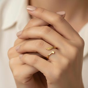 May include: A gold-colored ring with an open design. One end features a small wing, and the other end has a heart shape. The ring is worn on a finger, showcasing its delicate details and elegant style.