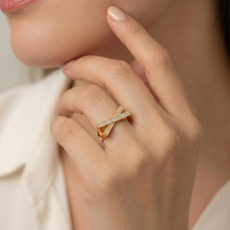 May include: A gold-colored ring with a crossed design. One band is encrusted with small, clear gemstones. The ring is worn on a finger, with the hand and part of a person's face visible in the image.