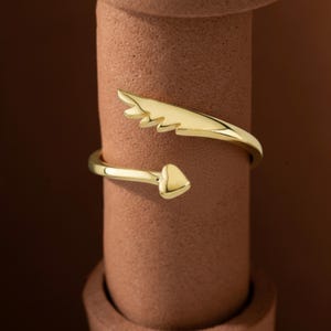 May include: A gold-colored ring with a wing-like design and a heart-shaped end. The ring wraps around the finger, creating a unique and stylish look. The ring is displayed on a brown cylindrical object.