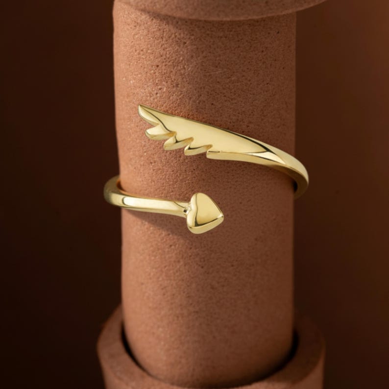 May include: A gold-colored ring with a wing-like design and a heart-shaped end. The ring wraps around the finger, creating a unique and stylish look. The ring is displayed on a brown cylindrical object.