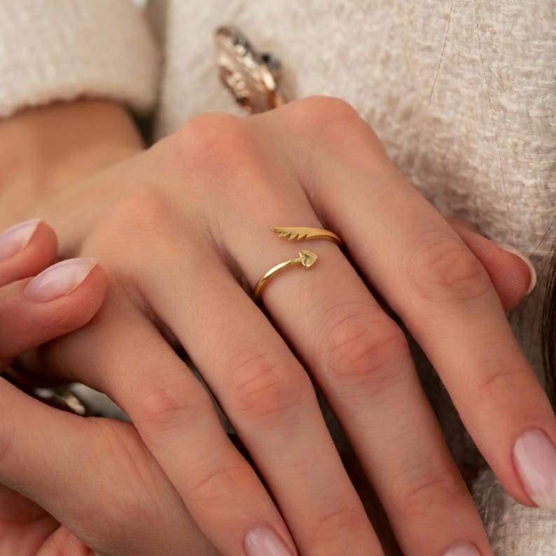 May include: A gold-colored ring with an arrow design. The ring features a small heart at the arrow's tip and a feather detail on the other end. The ring is worn on a finger.