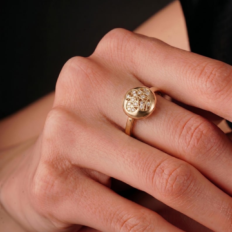 K&ouml;nnte beinhalten: Ein goldener Ring mit einer runden, strukturierten Oberfl&auml;che, die mit kleinen, funkelnden Edelsteinen verziert ist. Der Ring hat ein schlankes, poliertes Band, und die Edelsteine sind in einem Cluster-Muster angeordnet. Der Ring ist ein Schmuckst&uuml;ck.