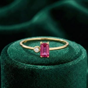 May include: A gold ring with a rectangular, bright pink gemstone and a small, round, clear stone. The ring is displayed on a dark green velvet surface, accentuating the ring's design.