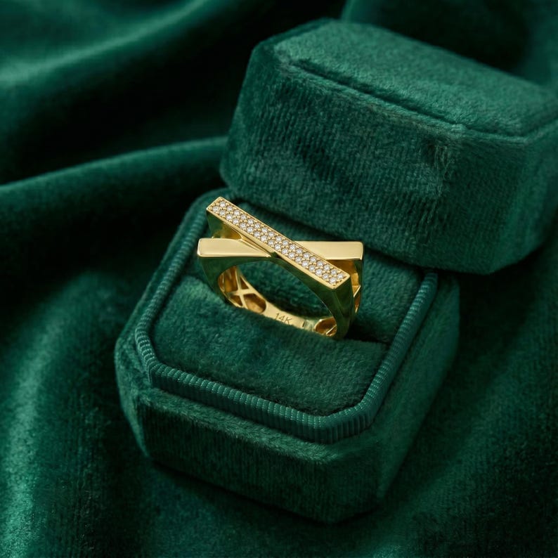 May include: A gold ring with a geometric design, featuring a band that crosses over another, adorned with small, sparkling stones. The ring is set in a dark green velvet jewelry box.