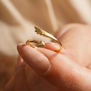 May include: A gold-colored ring with an open design. One end features a small heart, while the other has a wing-like shape. The ring is held up by a person's fingers, showcasing its delicate details and shiny finish.