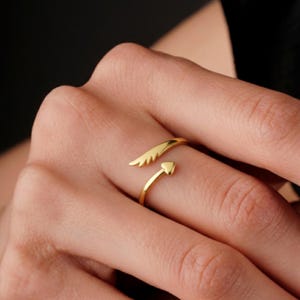 May include: A gold-colored ring with an open design. One end features a wing shape, while the other has a heart-shaped arrow. The ring is worn on a finger, showcasing its delicate details and shiny finish.
