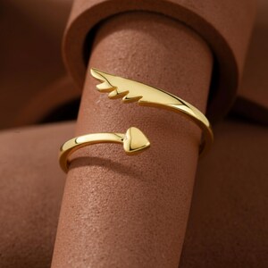 May include: A gold-colored ring with an open design. One end features a heart shape, while the other has a wing-like design. The ring is displayed on a brown surface, showcasing its shiny, reflective surface.