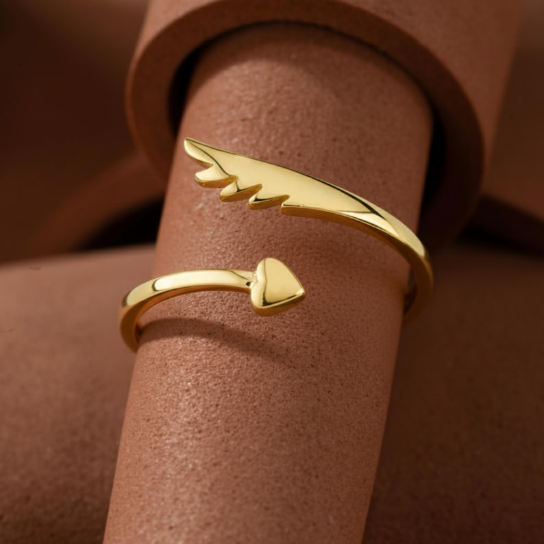 May include: A gold-colored ring with an open design. One end features a heart shape, while the other has a wing-like design. The ring is displayed on a brown surface, showcasing its shiny, reflective surface.