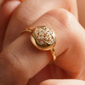 K&ouml;nnte beinhalten: Ein goldener Ring mit einem runden Gesicht, das mit einem geometrischen Muster aus kleinen Diamanten verziert ist. Der Ring hat einen warmen Goldton und wird am Finger getragen.