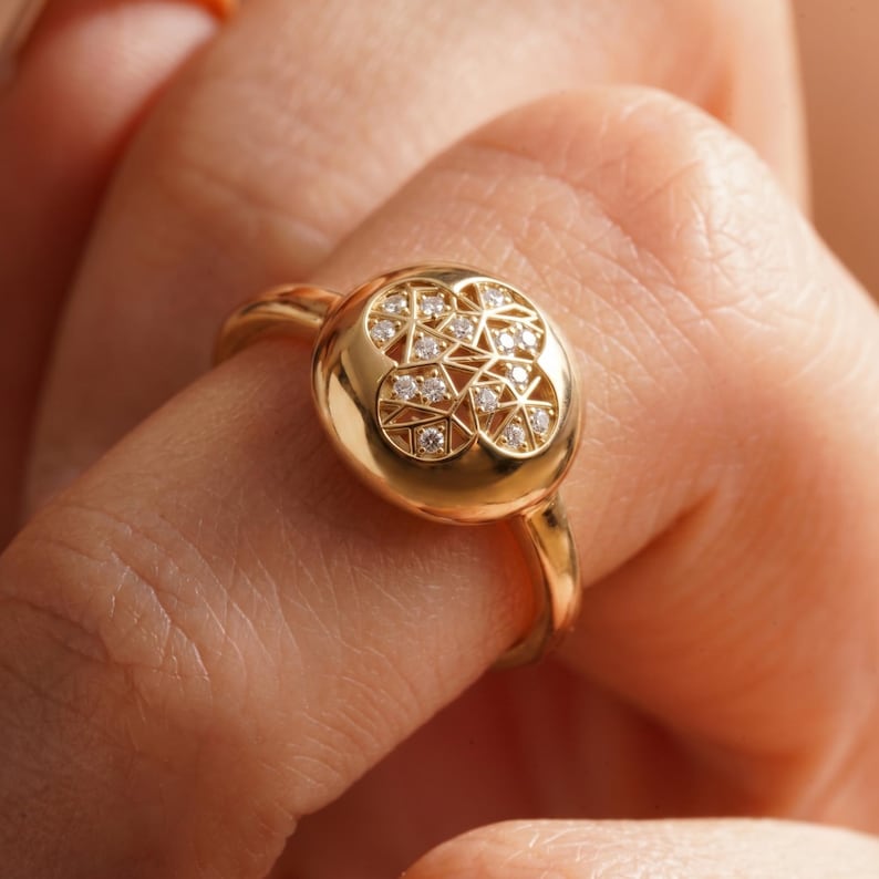 K&ouml;nnte beinhalten: Ein goldener Ring mit einem runden Gesicht, das mit einem geometrischen Muster aus kleinen Diamanten verziert ist. Der Ring hat einen warmen Goldton und wird am Finger getragen.