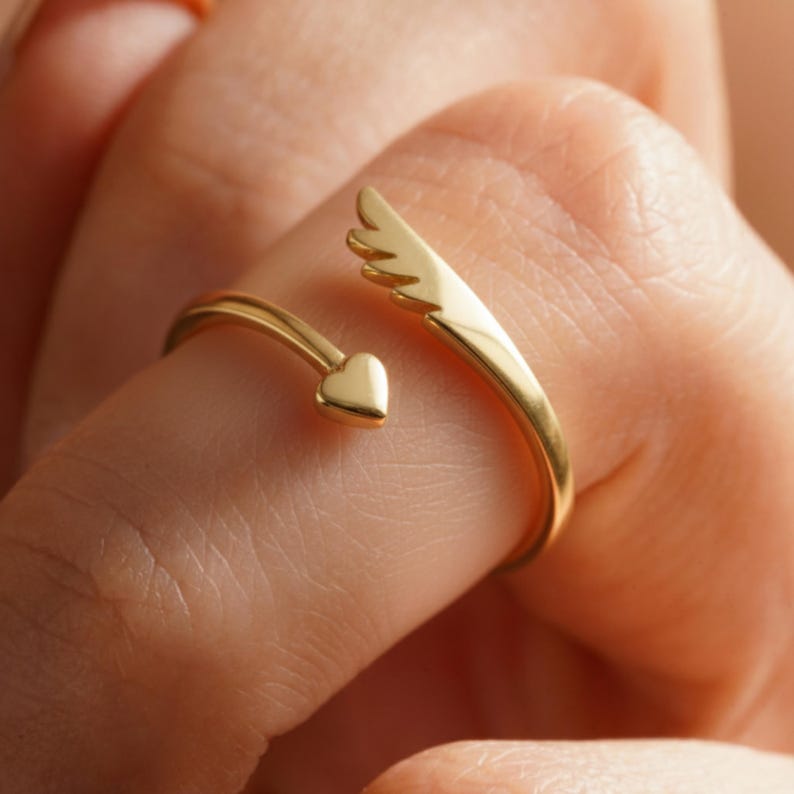 May include: A gold-colored ring with an open design. One end features a small heart, while the other has a wing shape. The ring is worn on a finger, showcasing its delicate details and shiny finish. This piece of jewelry is a romantic accessory.