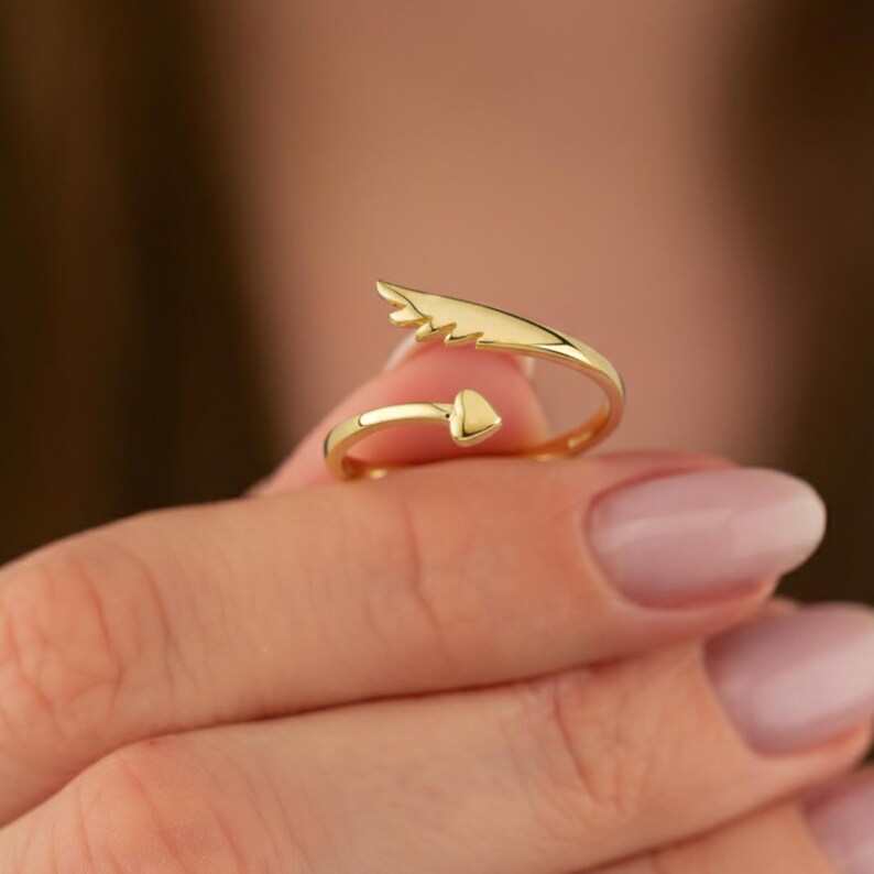 May include: A gold-colored ring with an open design. One end features a heart-shaped element, while the other has a stylized feather or wing shape. The ring is held up by a person's hand.