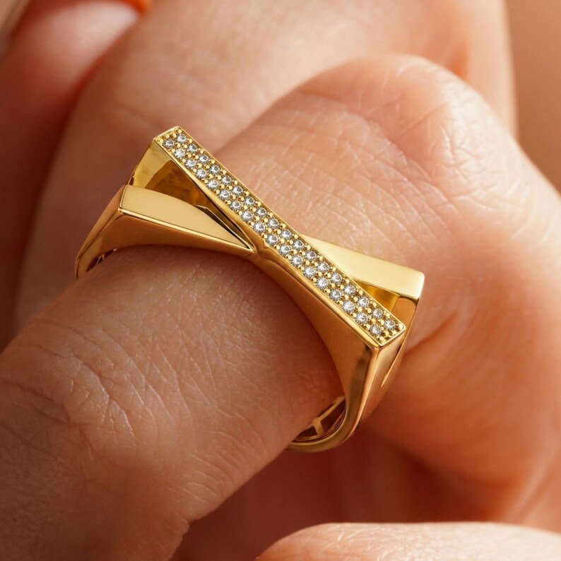 May include: A gold-colored ring with a geometric design. The ring features a band of small, sparkling stones set in a diagonal line across the top. The ring's design is modern and eye-catching.