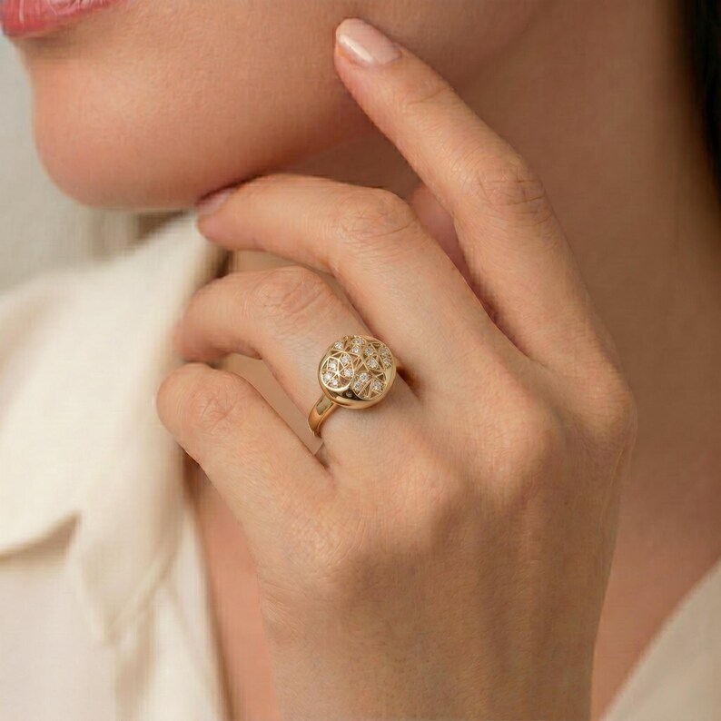 K&ouml;nnte beinhalten: Ein goldener Ring mit einer runden, strukturierten Oberfl&auml;che, besetzt mit kleinen, funkelnden Steinen. Der Ring befindet sich an einem Finger, die Hand in der N&auml;he des Gesichts einer Person. Der Ring hat ein schlichtes, poliertes Goldband.