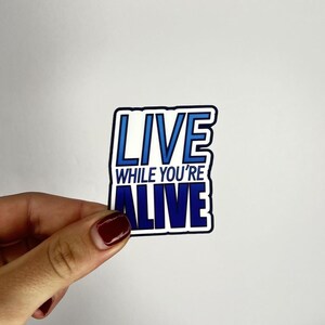 May include: Blue and white sticker with the text "LIVE WHILE YOU'RE ALIVE" in a bold, gradient font.