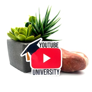 May include: A sticker with the YouTube logo, a red rectangle with a white play button, and the words "YOUTUBE UNIVERSITY". The sticker is next to a small, dark gray square pot with green plants and a reddish-brown stone.