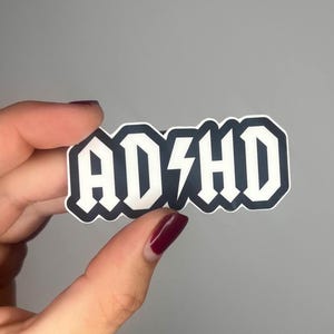 May include: White vinyl sticker with black lettering that reads "ADHD" in a bold, rock and roll font with a lightning bolt in between the "D" and "H".