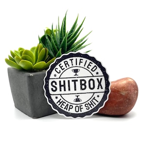 May include: A black and white sticker with the text "CERTIFIED SHITBOX" and "HEAP OF SHIT" is displayed. The sticker is next to a small succulent plant in a dark gray square pot and a reddish-brown stone.