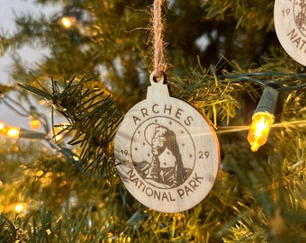May include: A wooden Christmas ornament with the text "Arches National Park" and a depiction of a rock formation. The ornament is hanging on a Christmas tree with lights.