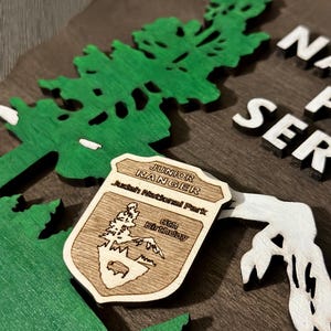 May include: A wooden sign with a green tree and the text "National Park Service" in white letters. A wooden badge with the text "Junior Ranger" and "Judah National Park" is attached to the sign. The badge also has the text "6th Birthday" on it.