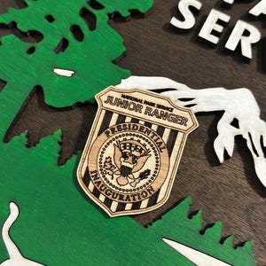 May include: A wooden badge with the text "National Park Service Junior Ranger" and "Presidential Inauguration" with a stylized eagle design. The badge is on a wooden background with a green tree and white mountain design.
