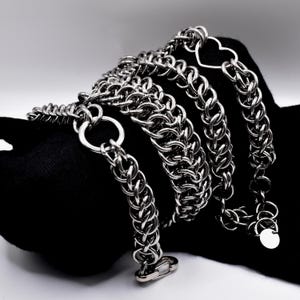 May include: A silver-colored chain necklace with a heart-shaped pendant and a circular tag. The chain is made of interlocking links and is displayed against a black background. The necklace has a clasp closure.