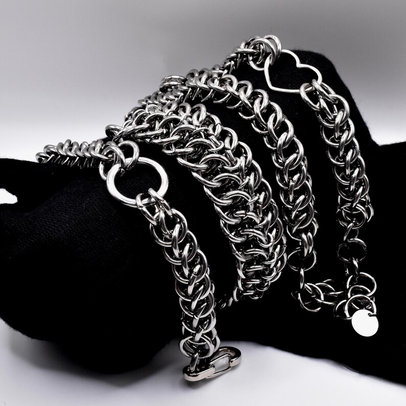 May include: A silver-colored chain necklace with a heart-shaped pendant and a circular tag. The chain is made of interlocking links and is displayed against a black background. The necklace has a clasp closure.
