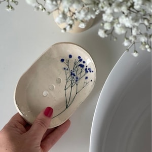 Può includere: Un portasapone in ceramica bianca con un design a fiori blu. Il portasapone ha tre fori di scarico.