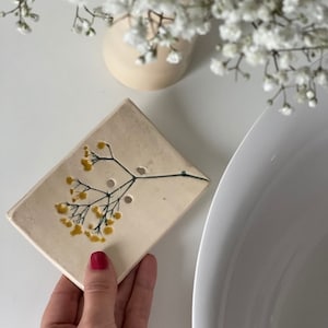 Puede incluir: Un jabonera rectangular de cerámica blanquecina con un diseño floral en amarillo y verde. La jabonera tiene tres orificios de drenaje. Un jarrón blanco con flores blancas está en el fondo.