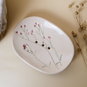 Könnte beinhalten: Eine weiße Keramik-Seifenschale mit Blumenmuster. Die Schale hat ein hellrosa und grünes Blumenmuster und drei Abflusslöcher.