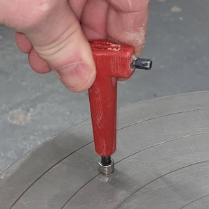 May include: A red plastic tool with a black metal tip is being used to centre a metal disc. The tool has the word "10V" printed on it.