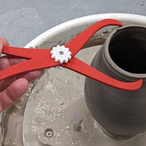 Könnte beinhalten: Ein rotes und weißes Töpfereigerät, das verwendet wird, um den Durchmesser eines Tontopfes zu messen. Das Gerät wird verwendet, um die Öffnung eines braunen Tontopfes auf einer Töpferscheibe zu messen.