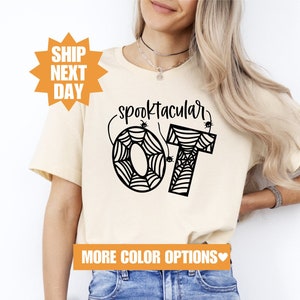 Può includere: Una maglietta beige con una grafica nera che dice "Spooktacular" con un design a ragnatela e le lettere "OT" al centro.