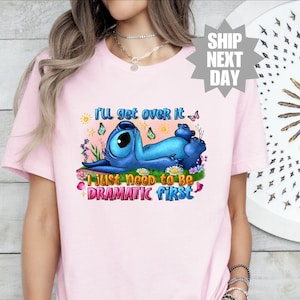 Op de afbeelding: Roze T-shirt met een blauw Stitch-personage uit Lilo & Stitch. Het shirt heeft de tekst "I'll get over it I just need to be dramatic first".