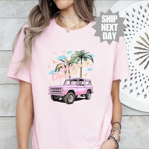 May include: Pink t-shirt featuring a graphic of a pink vintage Bronco driving through a sunset with palm trees and the text "Sunset Chaser".
