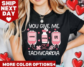 Op de afbeelding: Zwart T-shirt met een roze afbeelding van medische IV-zakken en de tekst "You Give Me Tachycardia". De IV-zakken zijn gelabeld met "Dopamine", "Atropine" en "EPI".