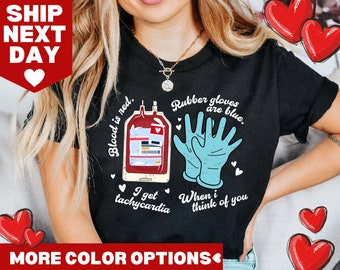 Op de afbeelding: Zwart T-shirt met een rode bloedzak en blauwe rubberen handschoenen als afbeelding. De tekst op het shirt luidt "Bloed is rood. Rubberen handschoenen zijn blauw. Ik krijg tachycardie wanneer ik aan je denk."