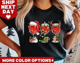 May include: Black t-shirt with a Christmas-themed design featuring three wine glasses with Santa hats, reindeer antlers, and a gingerbread man. The glasses are filled with red wine and decorated with Christmas lights and ornaments.