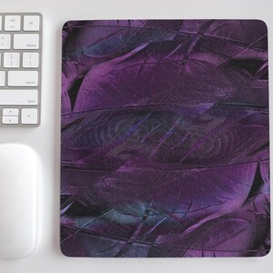 May include: A rectangular mousepad with a pattern of overlapping purple and teal feathers. The mousepad is next to a white computer mouse and a keyboard on a white surface.