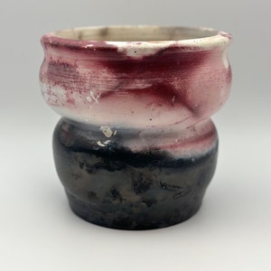 May include: A small, hand-thrown ceramic vase with a unique, abstract design. The vase is glazed in shades of white, pink, and black, with a textured finish.