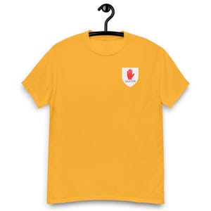 May include: Mustard yellow t-shirt with a small white and red Ulster hand symbol on the left chest. The t-shirt is hanging on a black hanger. The Ulster hand symbol is a red hand inside a white shield with the word "ULSTER" below it.