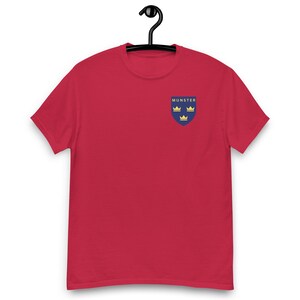 May include: A red t-shirt with a small crest on the left chest. The crest is a blue shield with the word "MUNSTER" at the top and three gold crowns. The t-shirt is hanging on a black hanger.